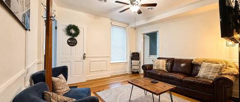 Un salon avec un ventilateur de plafond, un canapé en cuir marron, un fauteuil bleu et une table basse en bois.