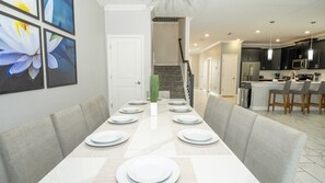 A dining area with a long table set for six, a staircase leading to an upper level, and a modern kitchen with dark cabinets and pendant lighting. - Near Disney 8BR Villa with Pool, Spa and Cinema (Kissimmee)