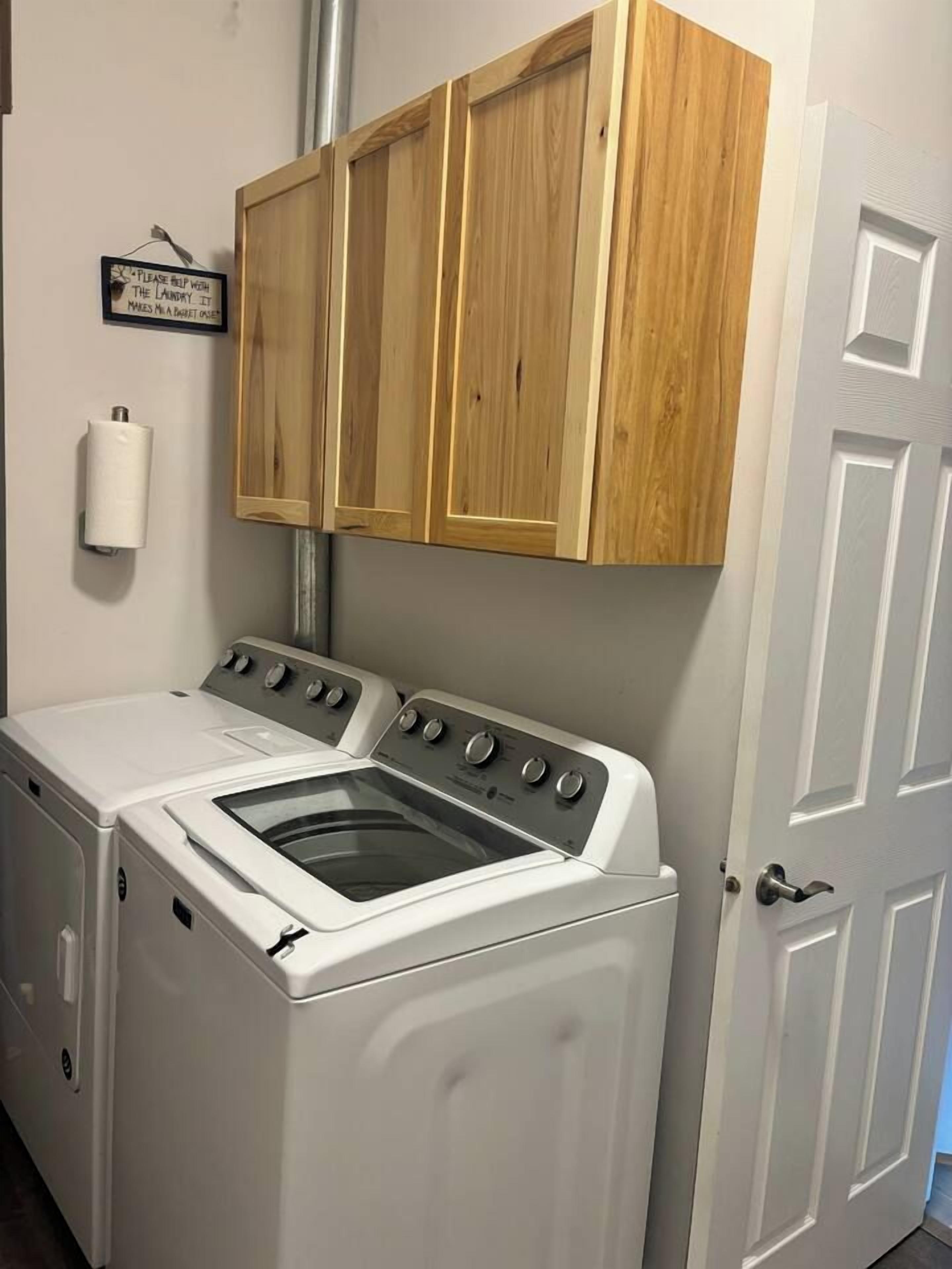A laundry room with a front-loading washer and dryer, wooden cabinets above, and a door to the right.