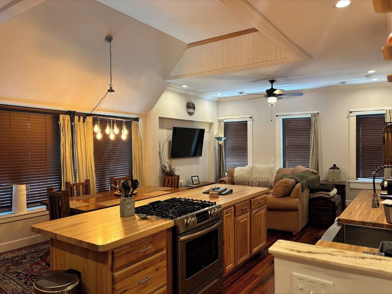 A compact kitchen with a central island, wooden cabinets, a built-in oven, and a stove. There is a dining area with a table and chairs, and a seating area with a sofa.