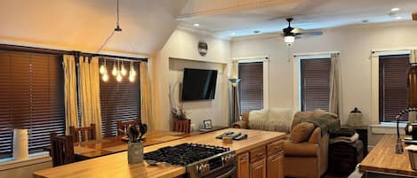 A compact kitchen with a central island, wooden cabinets, a built-in oven, and a stove. There is a dining area with a table and chairs, and a seating area with a sofa.