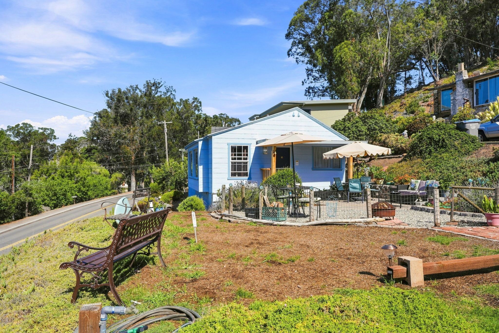 A blue house with a patio, surrounded by greenery and a road.