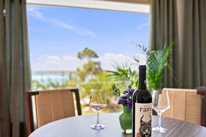 Ett bord med en vinflaska, två glas och en vas med blommor, placerat med utsikt över havet och grönskan.