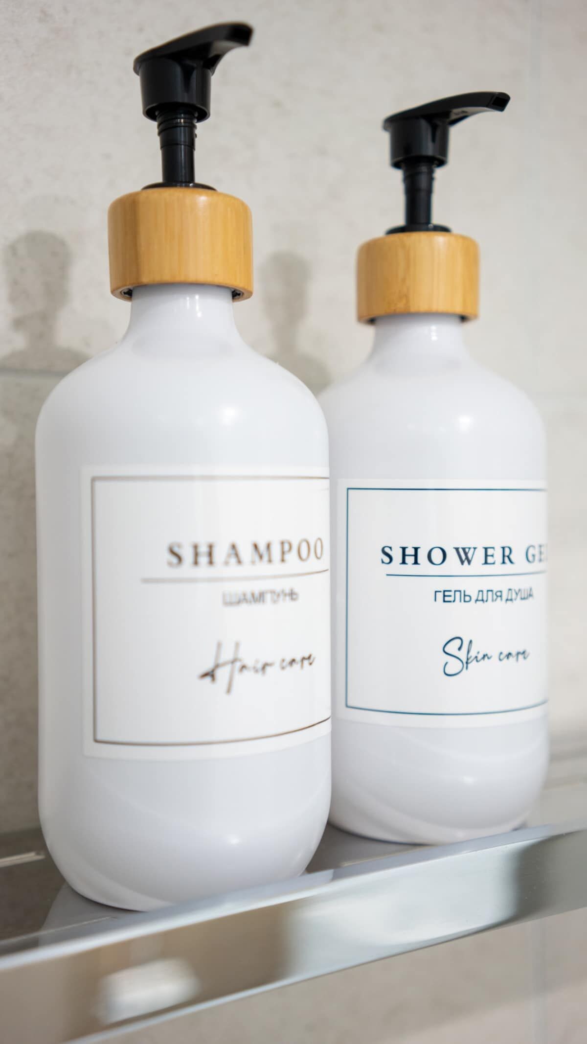 Two white bottles with wooden pump dispensers, one labeled 'SHAMPOO' and the other 'SHOWER GEL' with Cyrillic text.