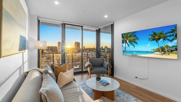 A modern living room with a sofa, armchairs, a coffee table, and a large TV screen displaying a tropical beach scene. - The Serenity | New Riverview Apt + Pool & Parking (Surfers Paradise)