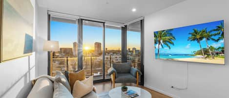 A modern living room with a sofa, armchairs, a coffee table, and a large TV screen displaying a tropical beach scene.