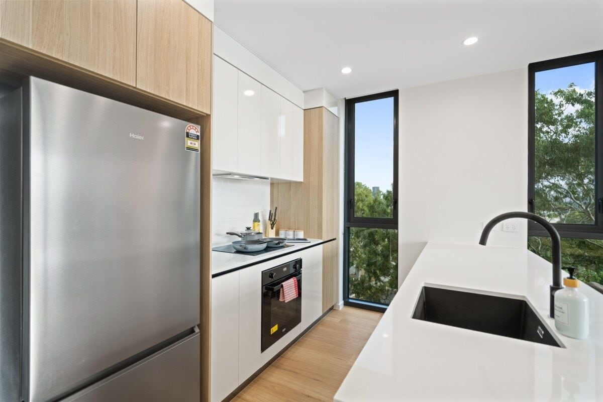 A modern kitchen with a stainless steel refrigerator, a built-in oven, and a large window providing a view of trees.