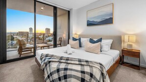 A modern hotel room with a large bed, a view of the cityscape, a painting on the wall, and a balcony with a chair. - The Serenity | New Riverview Apt + Pool & Parking (Surfers Paradise)