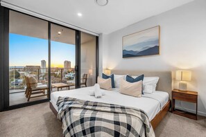 A modern hotel room with a large bed, a view of the cityscape, a painting on the wall, and a balcony with a chair. - The Serenity | New Riverview Apt + Pool & Parking (Surfers Paradise)