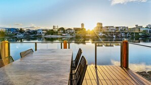A wooden deck with a table and chairs overlooking a calm body of water with buildings in the background. - The Serenity | New Riverview Apt + Pool & Parking (Surfers Paradise)