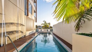 A swimming pool with a wooden deck, surrounded by a building with large windows and a palm tree. - The Serenity | New Riverview Apt + Pool & Parking (Surfers Paradise)