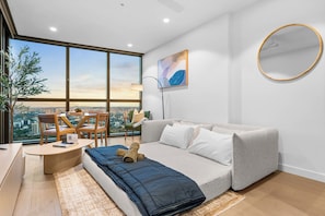 Ein modernes Wohnzimmer mit einer Couch, einem kleinen Tisch und einem Sessel. Ein großes Fenster bietet Blick auf die Stadt.