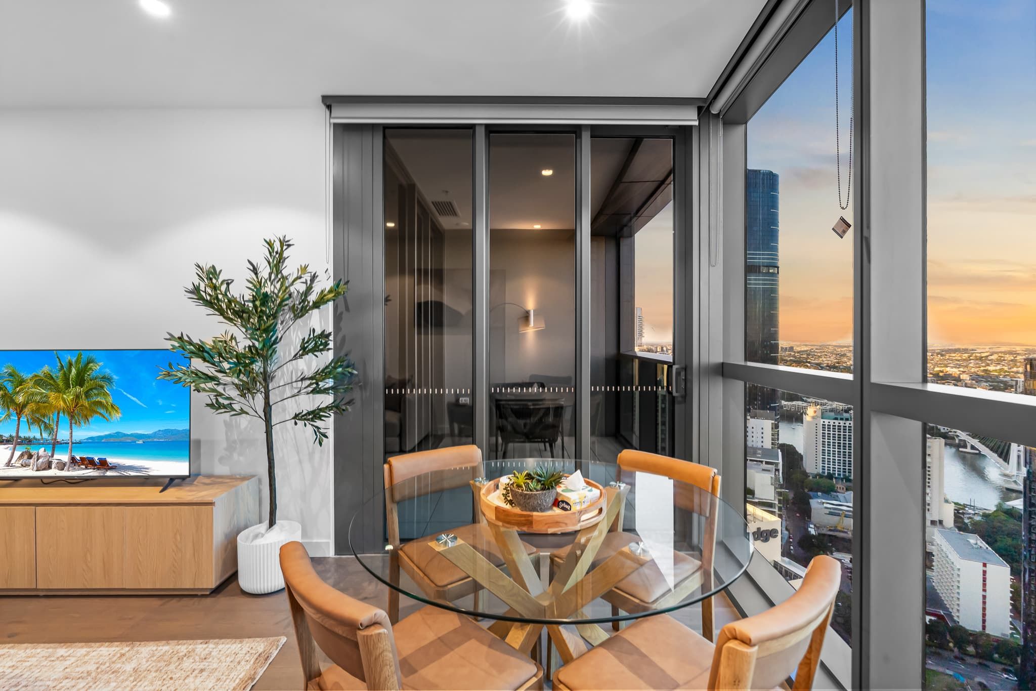 A modern interior with a dining area, a large window with a city view, and a television displaying a beach scene.