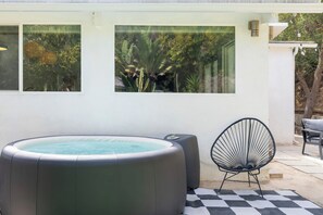 Un bain à remous, une chaise et un sol à carreaux dans un espace extérieur agrémenté d’un mur blanc et de fenêtres.