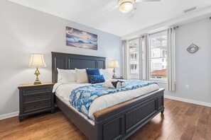 A bedroom with a dark wooden bed, two bedside tables with lamps, a ceiling fan, a large window with blinds, and a painting on the wall. - Sojourn 3BD2BA with Balcony, 3 Pools, Beach Access (Virginia Beach)