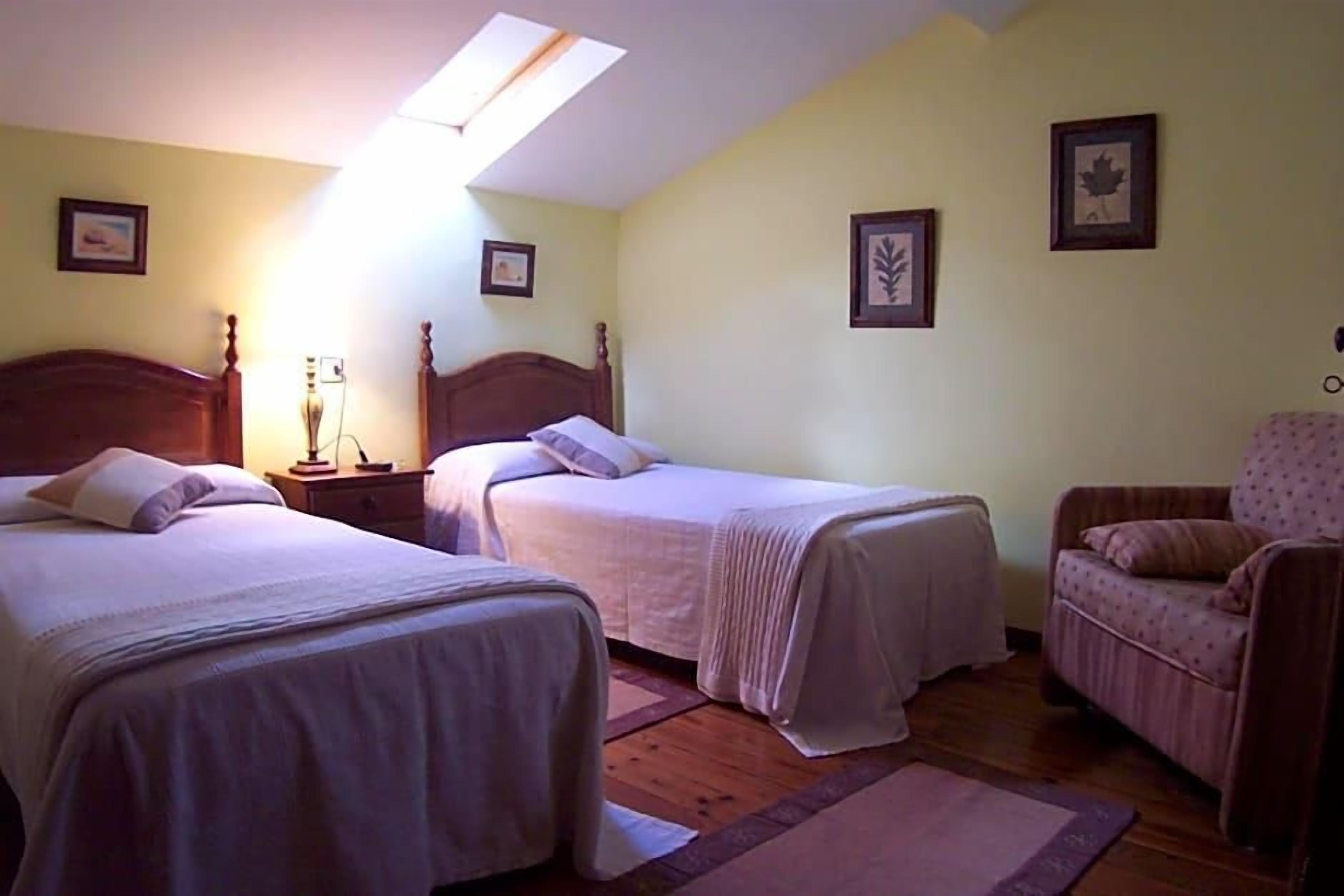 Un dormitorio con dos camas individuales, cabecera de madera, mesita de noche, una silla y un tragaluz.