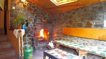 A stone-walled room with a fireplace, a wooden bench, a table with a checkered tablecloth, and a green metal container.