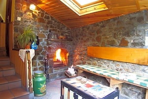 Una habitación con paredes de piedra, una chimenea, un banco de madera, una mesa con mantel a cuadros y un recipiente metálico verde.