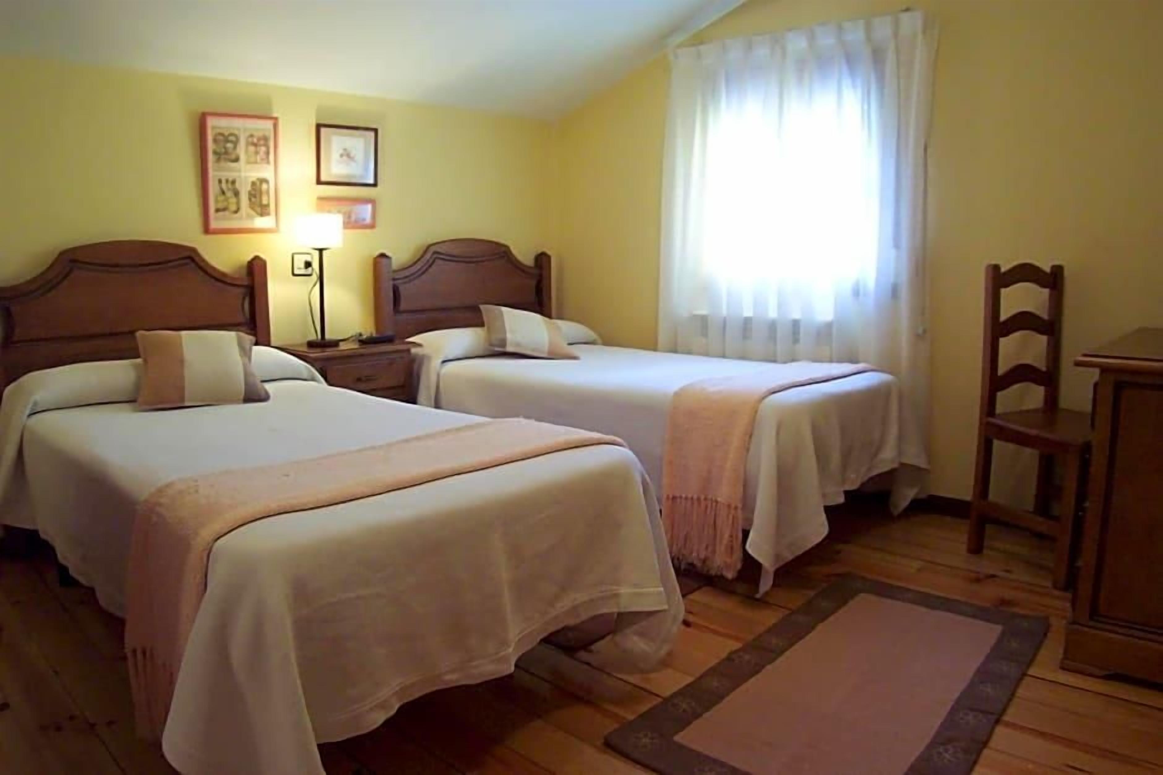A hotel room with two beds, a nightstand, a lamp, a chair, and a window with curtains.