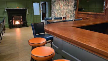 A bar area with a wooden counter, orange stools, and a fireplace.