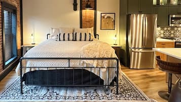 A bedroom with a large bed, a wooden floor, a brick wall, a kitchen with a refrigerator, and a window with blinds.