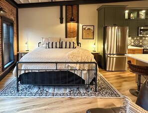 A bedroom with a large bed, a wooden floor, a brick wall, a kitchen with a refrigerator, and a window with blinds.