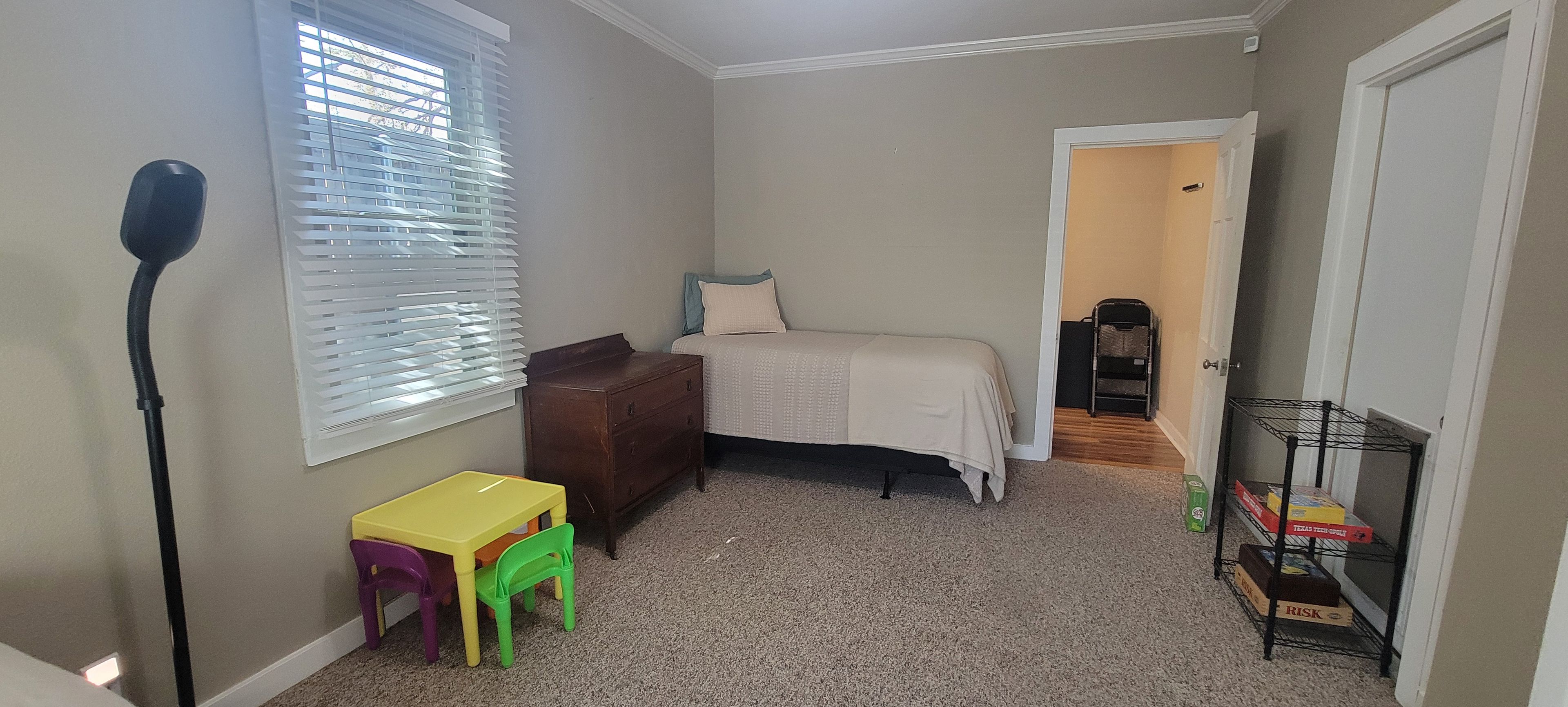 Een slaapkamer met een bed, een kaptafel, een bureau met stoelen, een radiator, een radiatoromsluiting, een deur en een plank met dozen.