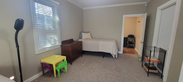 A bedroom with a bed, a dresser, a desk with chairs, a radiator, a radiator cover, a door, and a shelf with boxes.