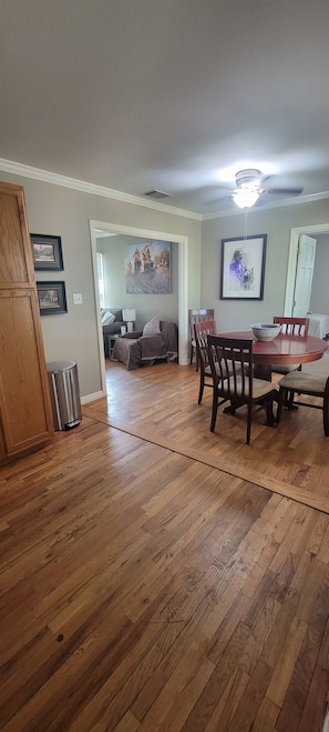 A dining area with a wooden table and chairs, a ceiling fan, and a hallway leading to a living room.