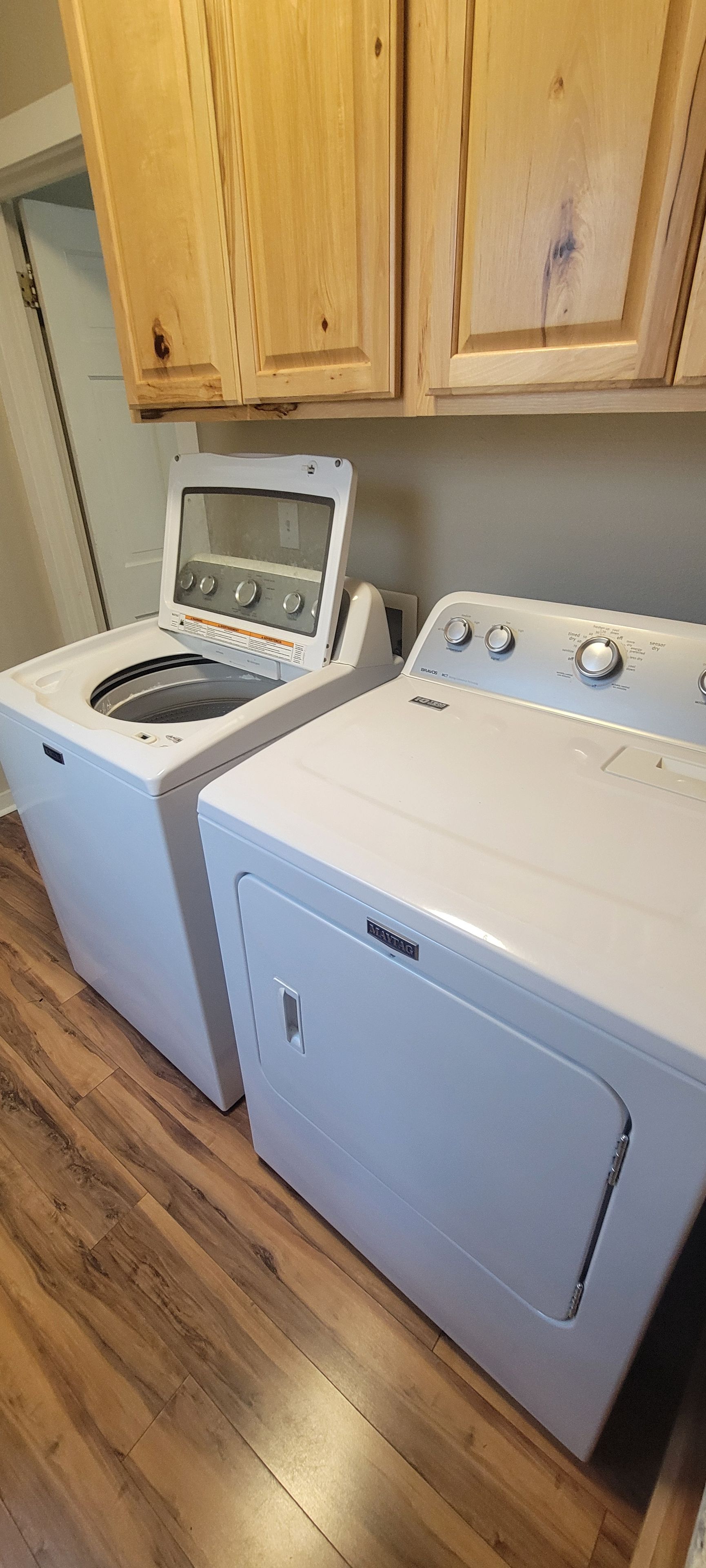 A laundry area with a front-loading washer and dryer, wooden cabinets above, and a wooden floor.
