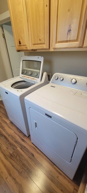 A laundry area with a front-loading washer and dryer, wooden cabinets above, and a wooden floor.