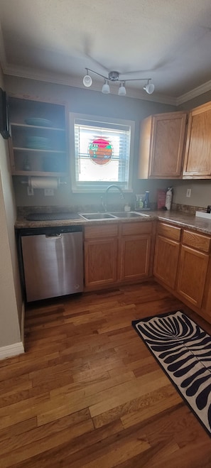 A compact kitchen with wooden cabinets, a stainless steel dishwasher, a window with blinds, and a zebra-patterned rug on the floor.