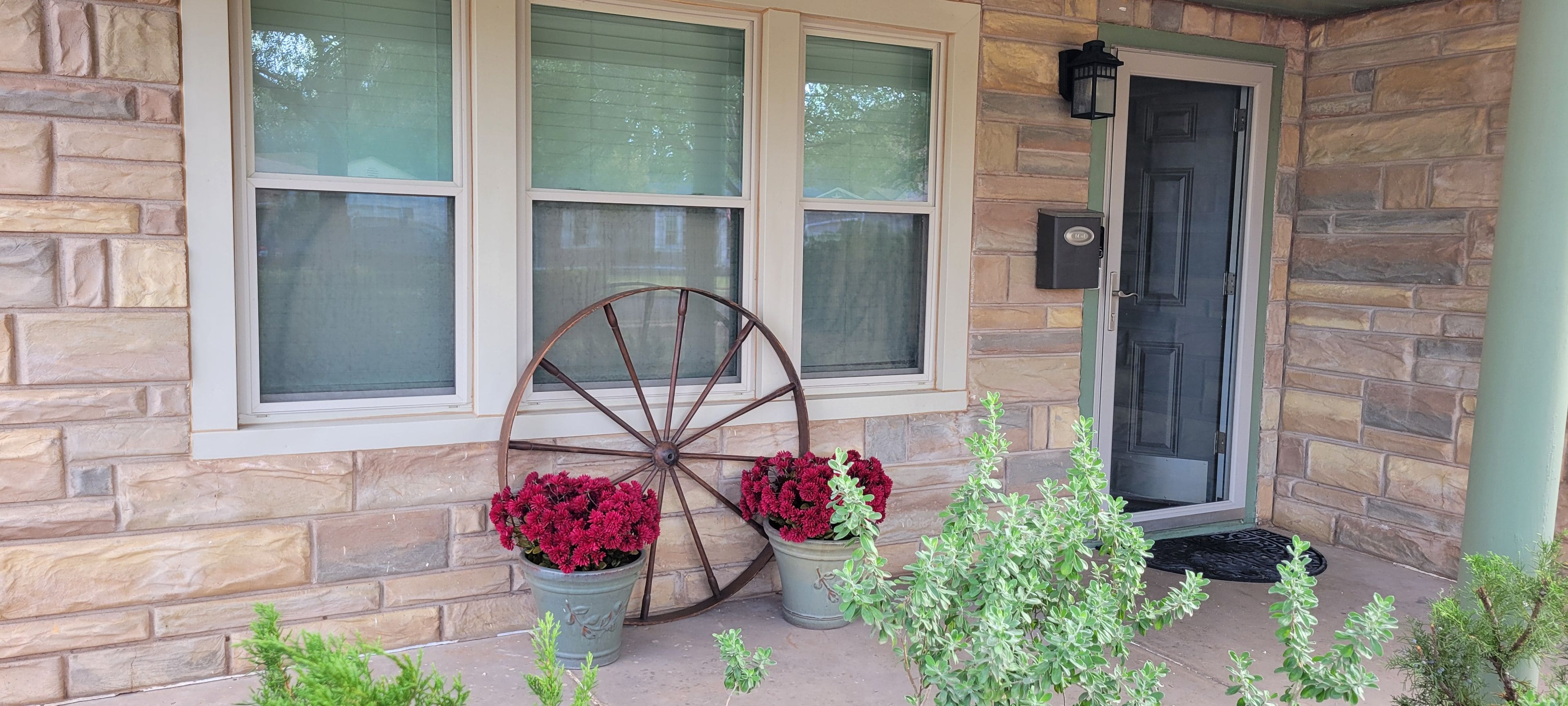 A stone house with a front door, two potted plants with red flowers, and a large wooden wheel.