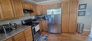 A kitchen with wooden cabinets, a stainless steel refrigerator, a microwave, a stove, and a trash can.