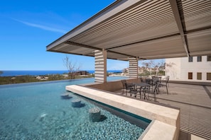 Una piscina infinita azul cristalina con vistas al océano, un área de estar techada al aire libre con mesas y sillas, y un diseño arquitectónico moderno.