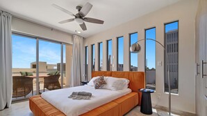A modern hotel room with a large bed, a ceiling fan, a floor lamp, and a view of the city through large windows. - Vista Añuiti: 2BR w/ Spectacular Ocean Views (Cabo San Lucas)