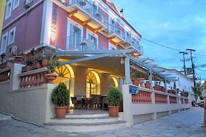 A hotel with a covered outdoor seating area, multiple balconies, and potted plants. - Seven Brothers Htl (Poros)