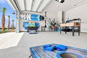A spacious room with a high ceiling, a ceiling fan, and a large window allowing natural light. There are bicycles and a surfboard mounted on the wall, and a table with a blue cushion.