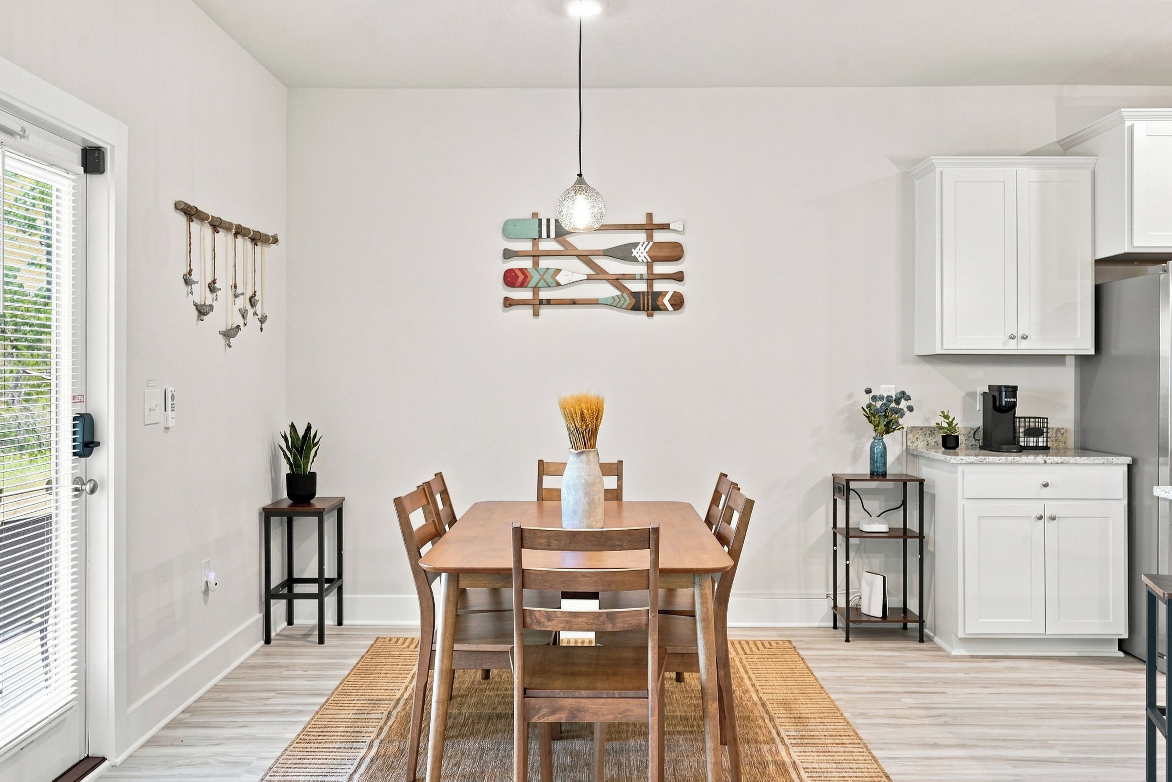 Uma sala de jantar com mesa de madeira, cadeiras, luminária pendente e cozinha ao fundo.