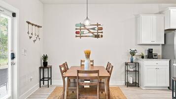 Uma sala de jantar com mesa de madeira, cadeiras, luminária pendente e cozinha ao fundo.
