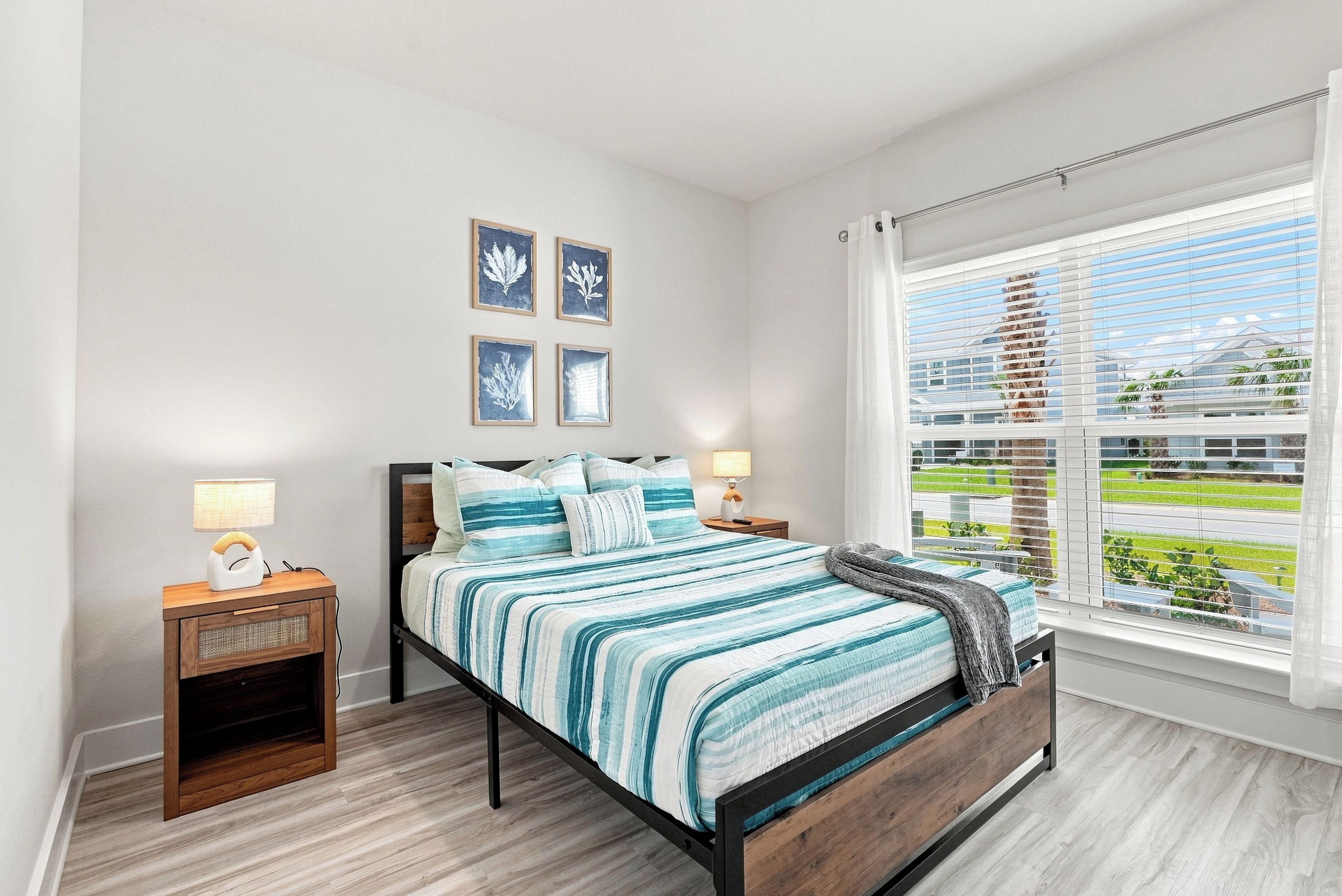 A bedroom with a bed, bedside tables, a window with blinds, and wall-mounted artwork.