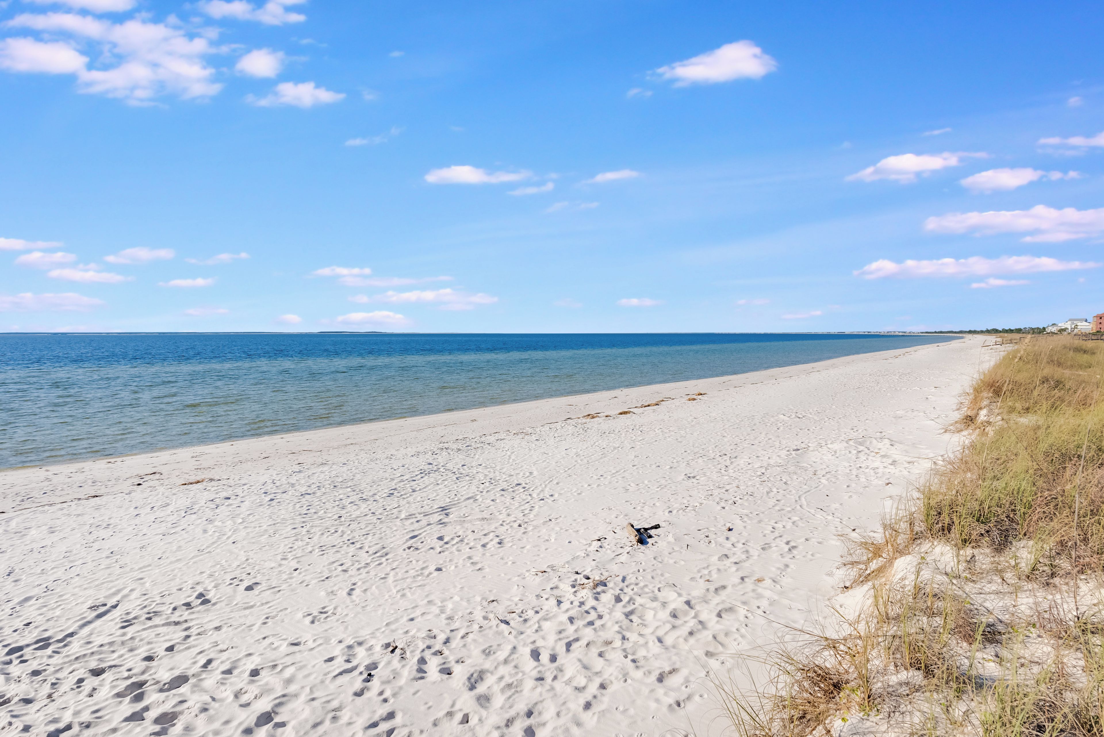 A wide sandy beach with footprints leading to the water, a clear sky, and distant buildings.