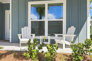 Un patio couvert, agrémenté de chaises Adirondack blanches et d’une table, entouré de petites plantes et d’un paillis d’aiguilles de pin.