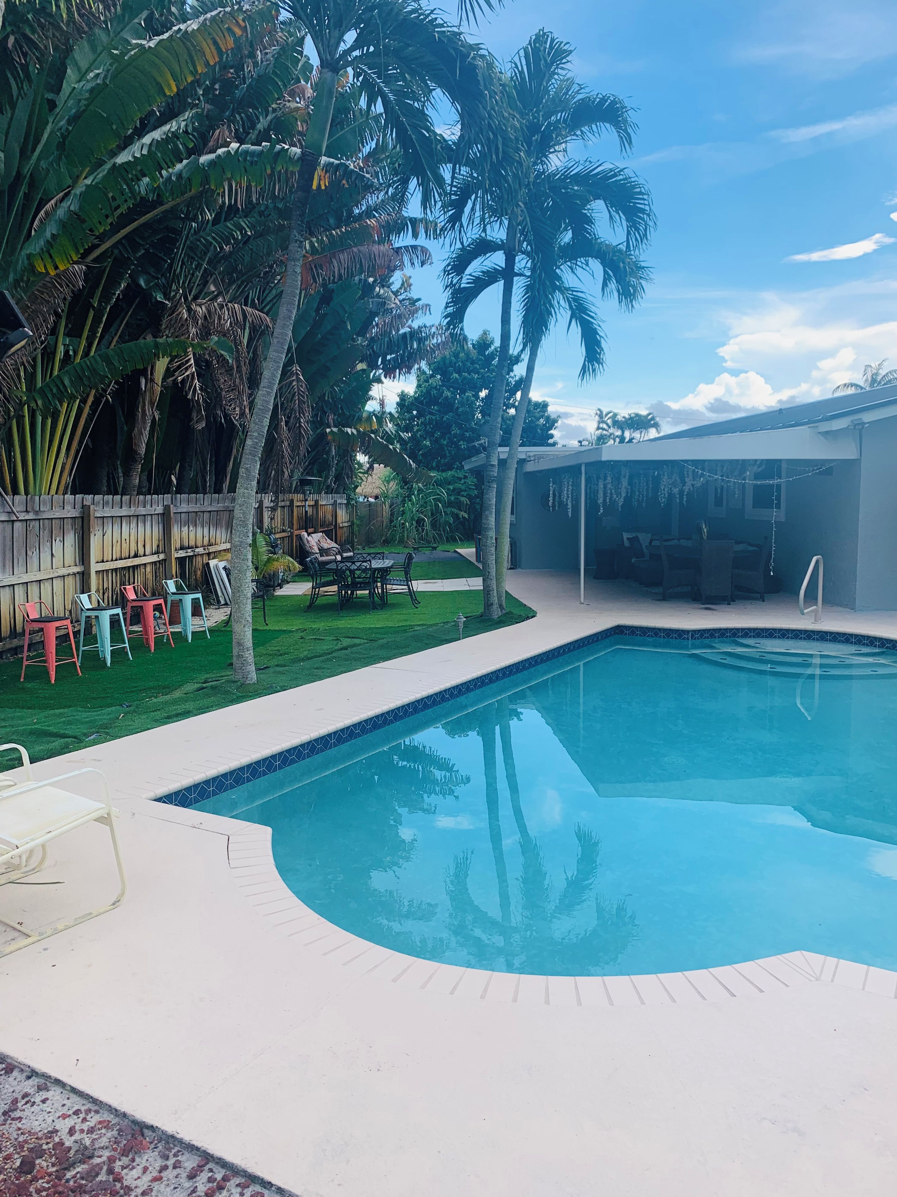 Una piscina con borde de azulejos, rodeada de césped, palmeras y una valla de madera.