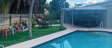 Una piscina con borde de azulejos, rodeada de césped, palmeras y una valla de madera.