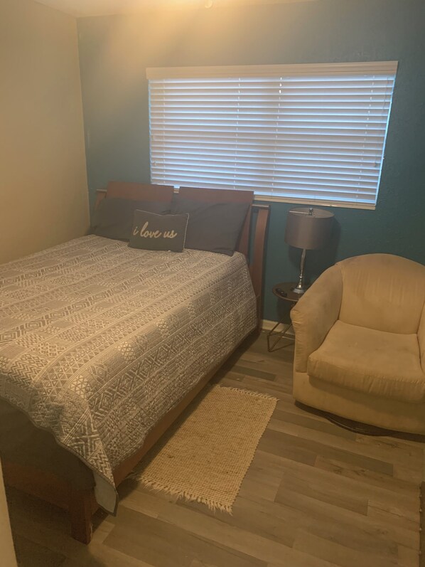 Un dormitorio con cama, una silla, un ventanal con persianas y una lámpara.