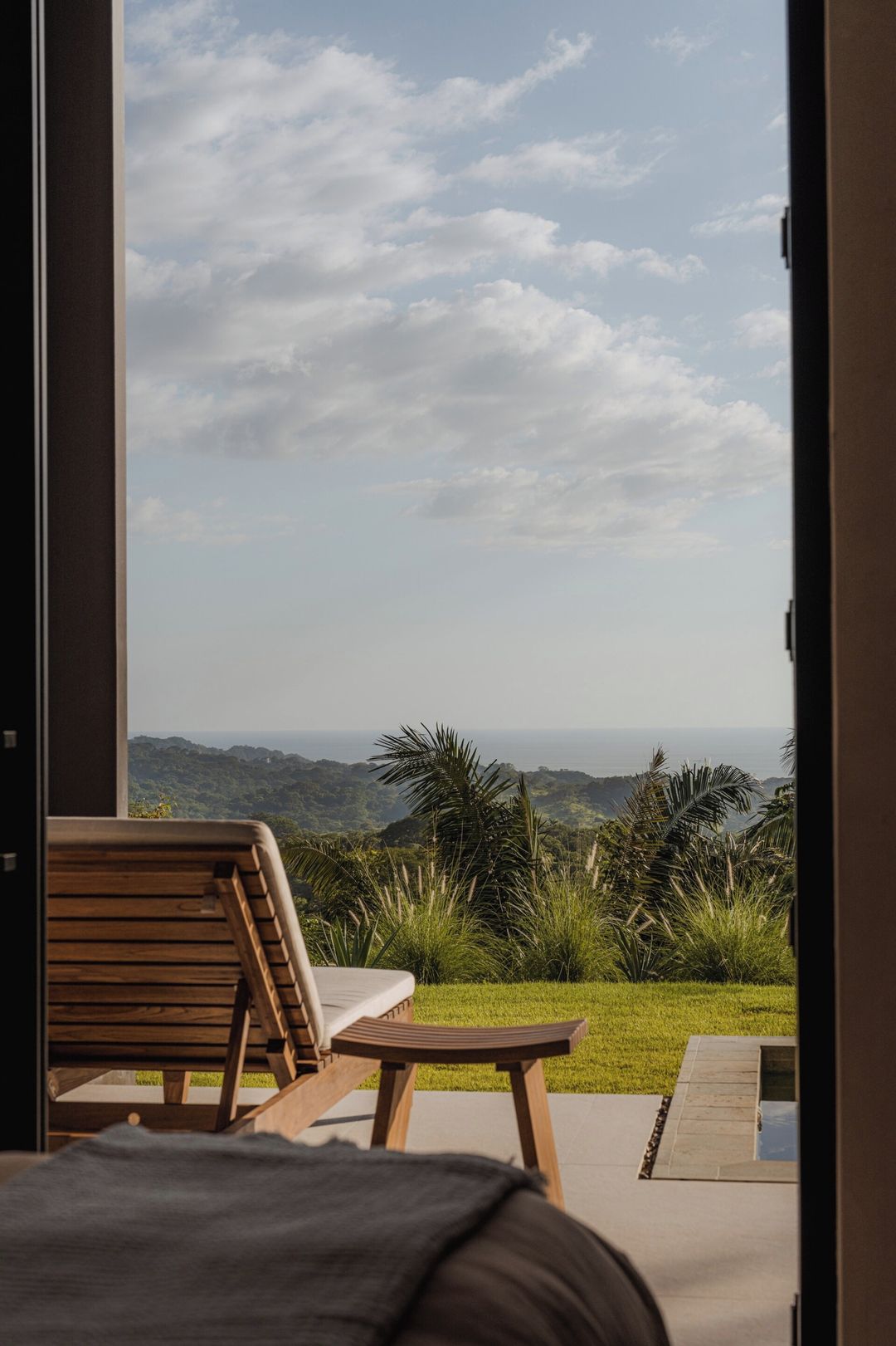 Una habitación con una cama, una silla de madera y una pequeña mesa. Se aprecia una vista de un paisaje exuberante con árboles y un horizonte lejano.