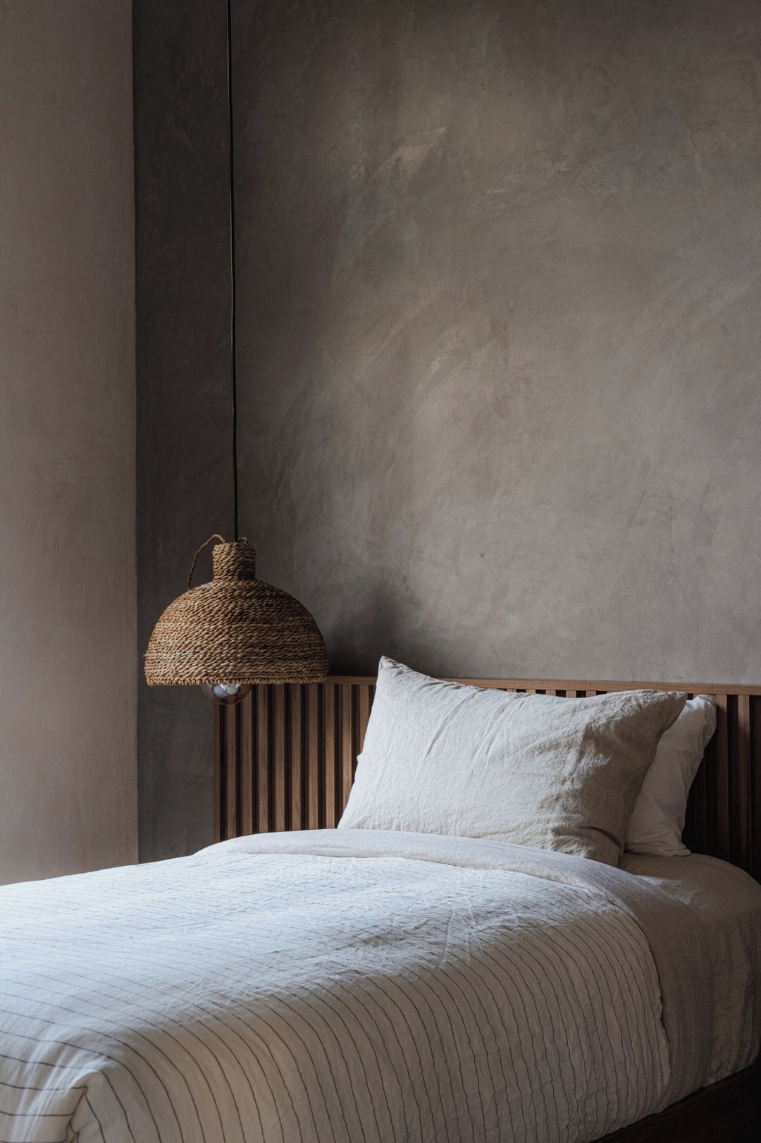 Une chambre à coucher avec un lit, un luminaire suspendu en fibres tressées et un mur uni.