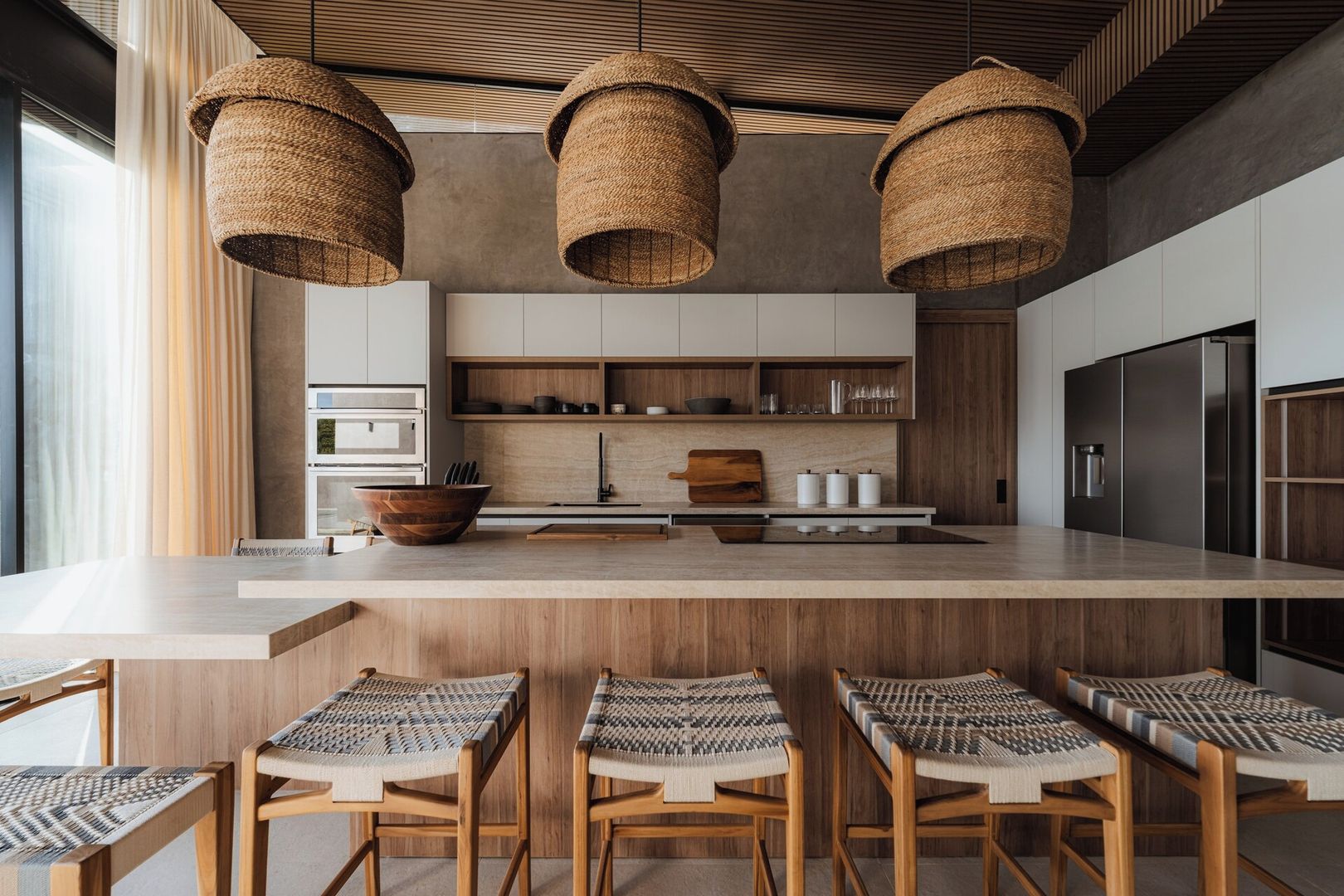A modern kitchen with a central island, hanging woven light fixtures, and wooden chairs with woven seats.