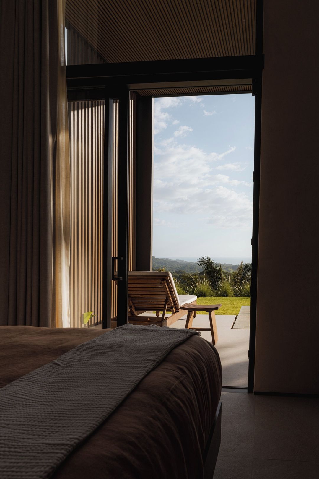 Un dormitorio con una cama grande, una silla de madera y vistas a un paisaje verde a través de una puerta abierta.